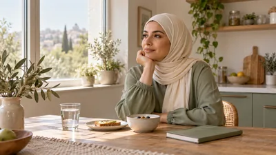 امرأة بملابس محتشمة قرب نافذة أمام وجبة صغيرة وكوب ماء ودفتر مغلق
